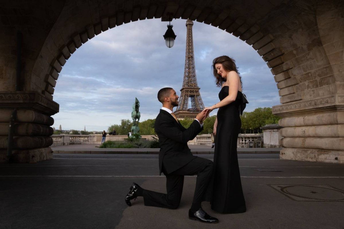 Couple élégant lors d’une demande en mariage sous un pont face à la Tour Eiffel à Paris, l’homme à genoux tenant la main de sa partenaire dans un moment romantique.
